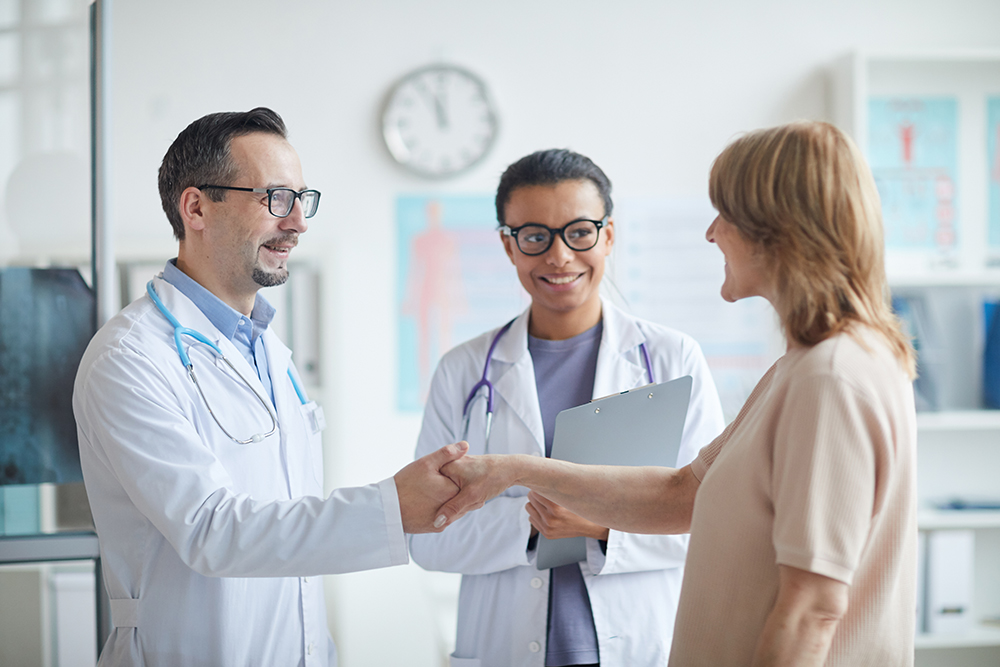 Mature male doctor shaking hands to mature woman with nurse standing near by them and smiling they cooperating at clinic